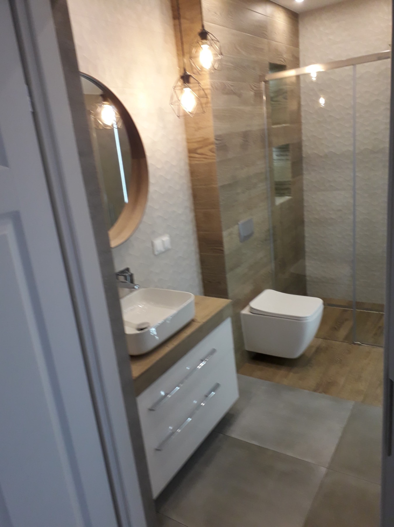 Nowoczesna łazienka z okrągłym lustrem, wiszącą szafką pod umywalką, toaletą podwieszaną i kabiną prysznicową z geometrycznymi płytkami na ścianach, oświetlona dekoracyjnymi lampami wiszącymi.