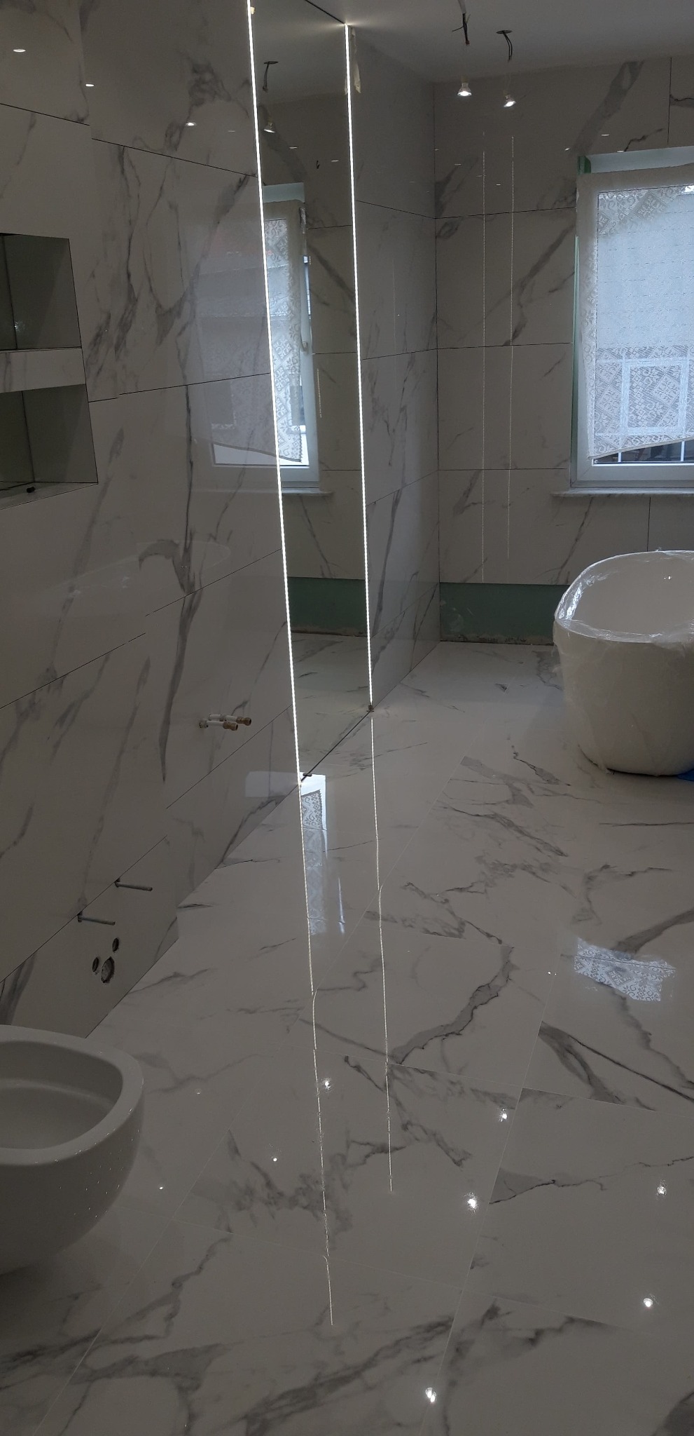 Nowoczesna łazienka w trakcie prac wykończeniowych: białe marmurowe płytki na ścianach i podłodze, pionowe pasy oświetlenia LED, częściowo zakryta folią ochronną wanna wolnostojąca.