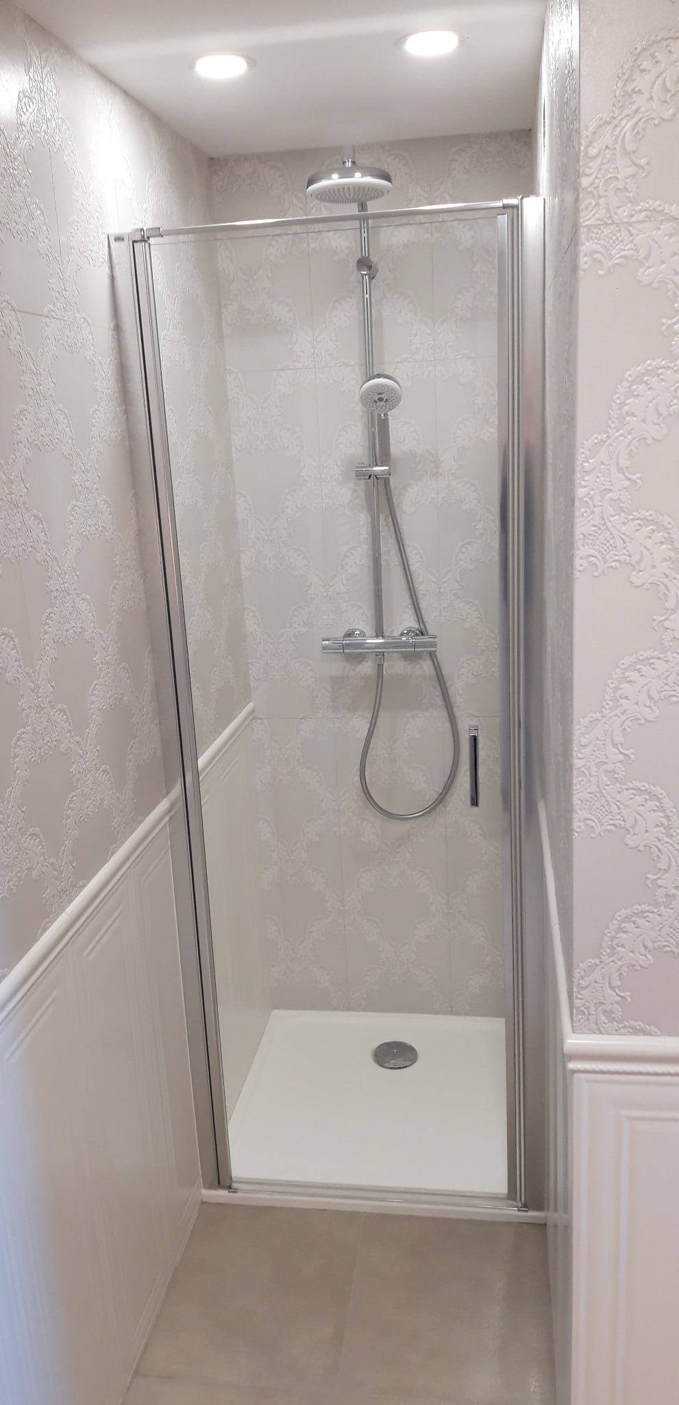 Nowoczesna kabina prysznicowa z deszczownicą i termostatem, otoczona białymi płytkami z delikatnym wzorem i listwami dekoracyjnymi.