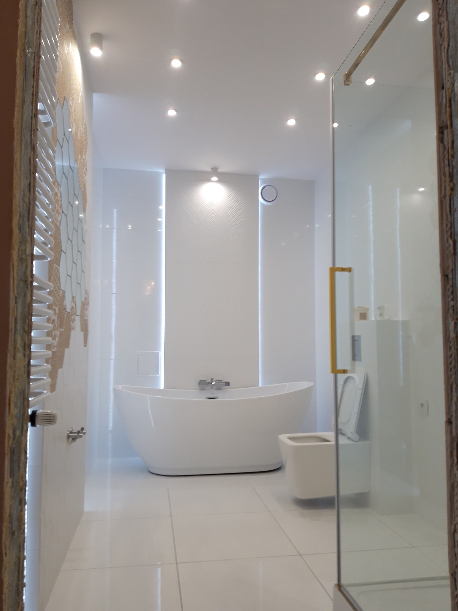 Nowoczesna łazienka z wolnostojącą białą wanną, podświetlanymi wnękami, szklaną kabiną prysznicową z złotym uchwytem i grzejnikiem drabinkowym z dekoracyjną ścianą w tle.