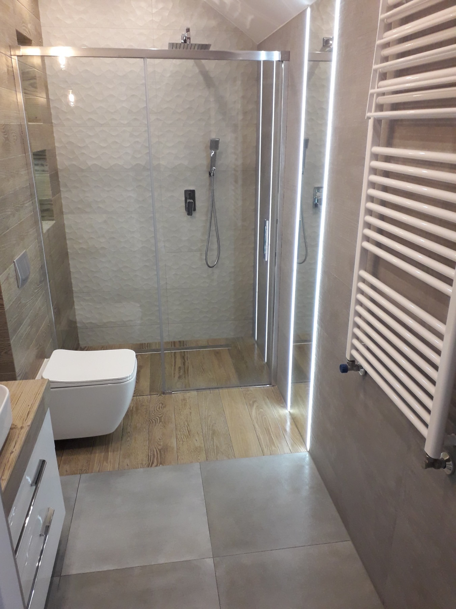 Nowoczesna łazienka z kabiną prysznicową z przesuwnymi drzwiami, podwieszaną toaletą, grzejnikiem drabinkowym i kombinacją płytek imitujących drewno oraz szarych, gładkich płytek podłogowych.