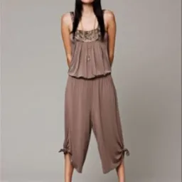 Kobieta w brązowym kombinezonie z szerokimi nogawkami do kolan, z wiązaniami na końcach, stojąca na szpilkach na szarym tle.