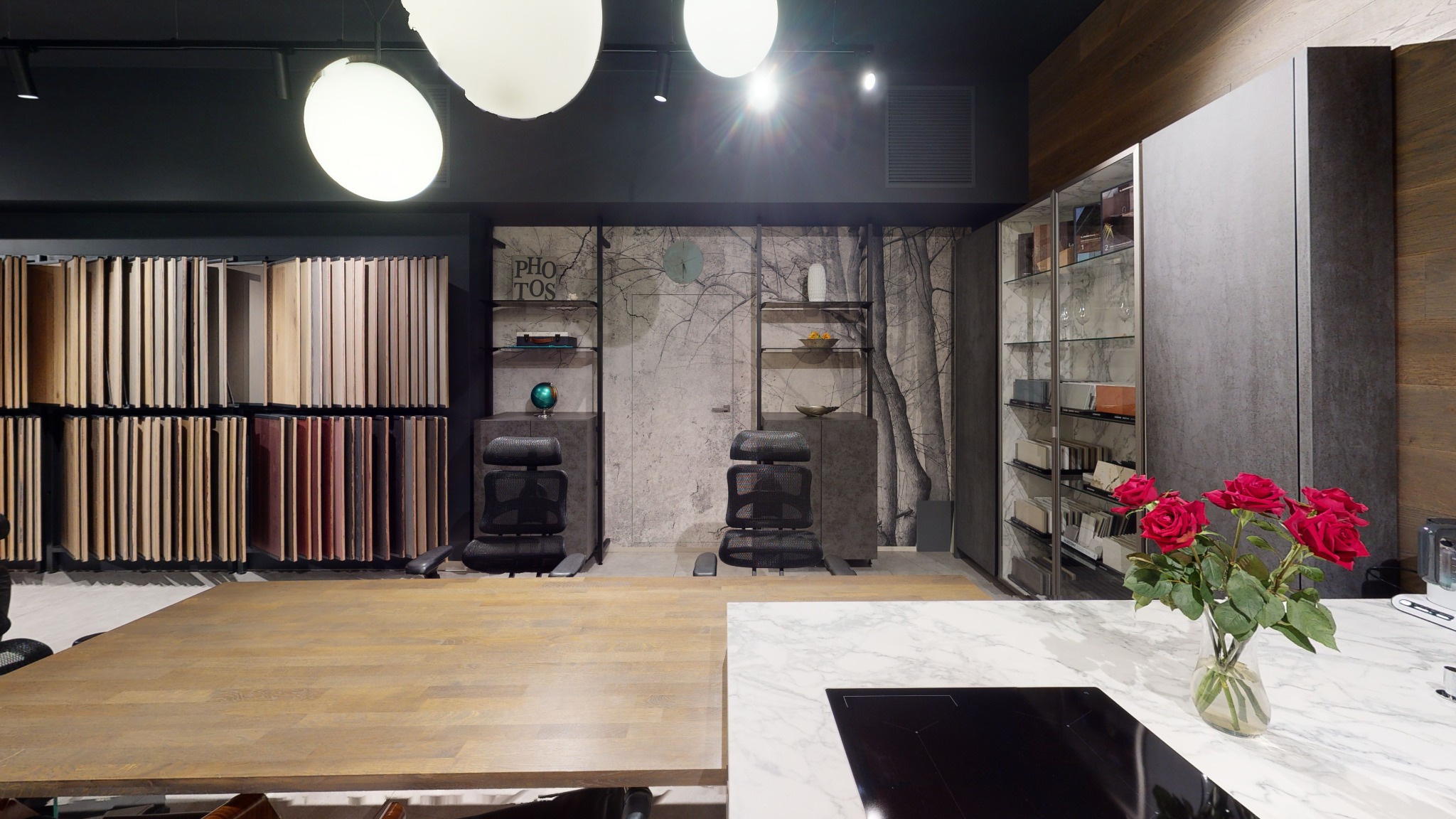 Wnętrze biura projektowego z ekspozycją próbek podłóg, mebli i okładzin ściennych, oświetlone kulistymi lampami sufitowymi, z bukietem czerwonych róż na pierwszym planie.