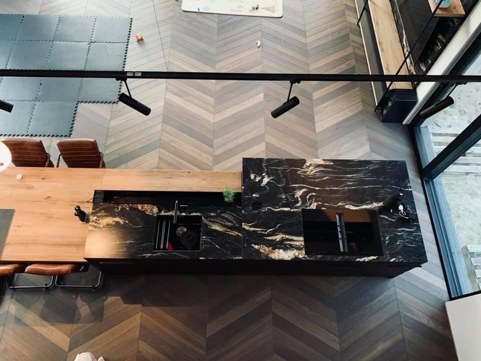 Widok z góry na kuchnię z parkietem ułożonym we wzór jodełki, czarny blat z marmurowym wzorem i drewniany stół z krzesłami, oświetlenie szynowe.