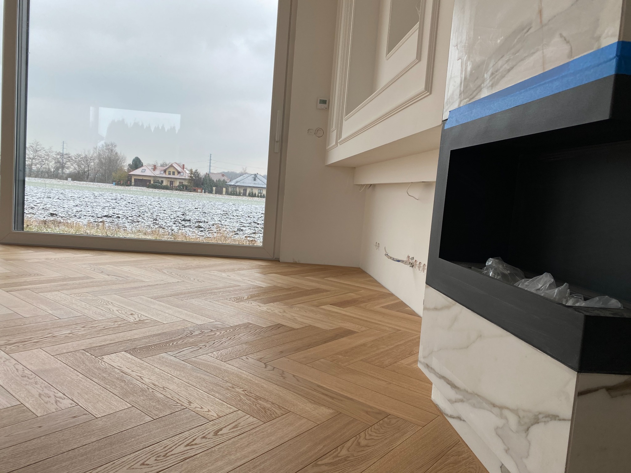 Jasny parkiet ułożony we wzór jodełki francuskiej w nowym wnętrzu z widokiem na zaśnieżone pole i domy za oknem, obudowa kominka zabezpieczona taśmą malarską.