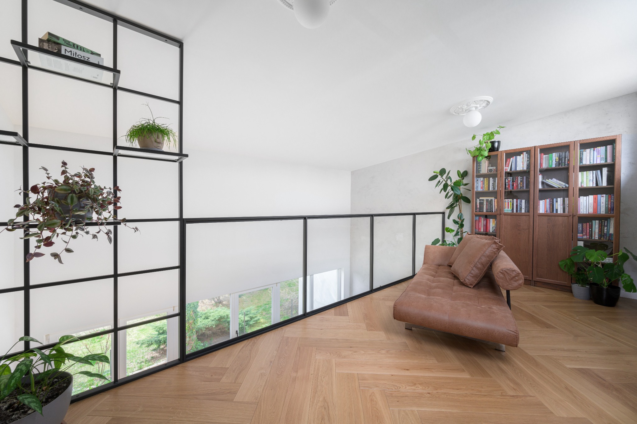 Salon na antresoli z parkietem ułożonym we wzór jodełki, brązową kanapą, regałem bibliotecznym i ścianką działową z czarnymi ramami i mlecznym szkłem.