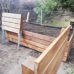 Anonim - Podwyższona grządka warzywna z drewnianą obudową na metalowych nogach, wypełniona ziemią, widoczna siatka ogrodzeniowa w tle.