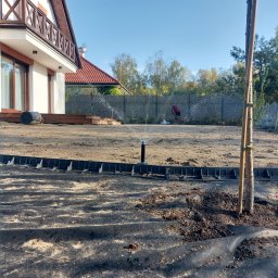 Anonim - Nowo założony trawnik z systemem nawadniania w trakcie testowania, widoczna czarna agrotkanina, obrzeża trawnikowe, świeżo posadzone drzewko podparte palikami i osoba pracująca w oddali.