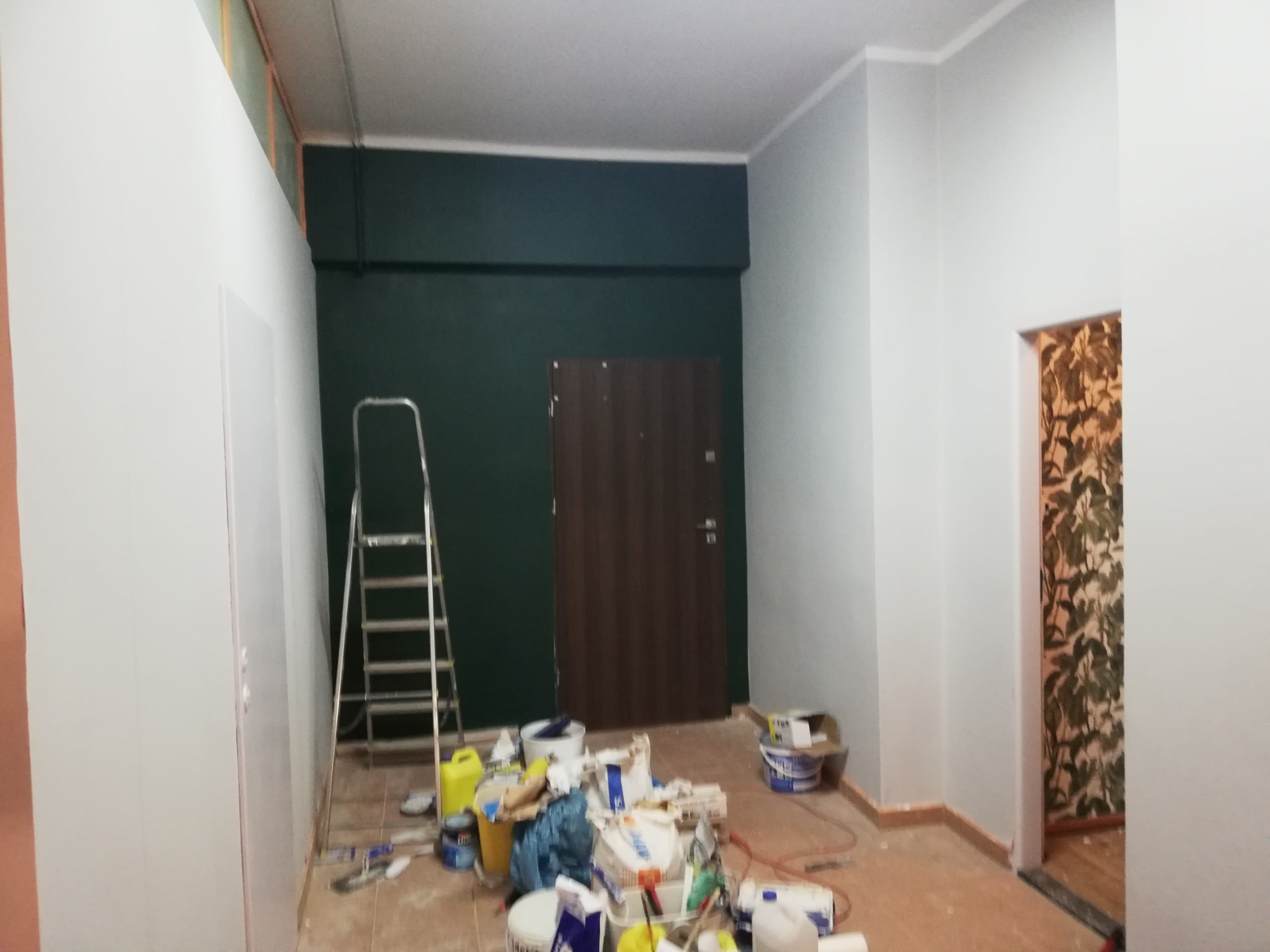 Pokój w trakcie malowania: ściany częściowo pomalowane na szaro i ciemnozielono, widoczna drabina, puszki z farbą i narzędzia malarskie na podłodze.