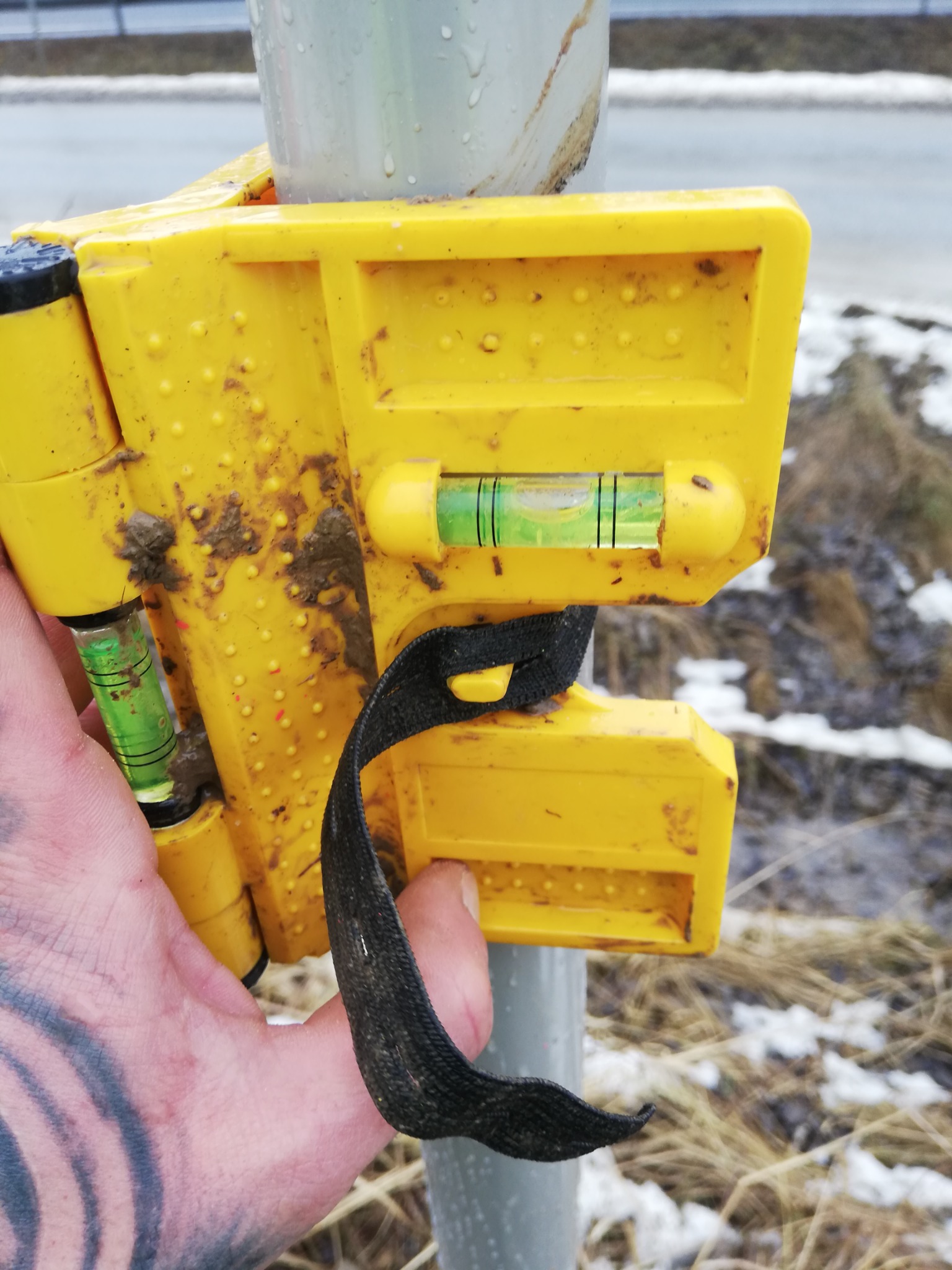 Żółta poziomica z paskiem, zabrudzona ziemią, przymocowana do szarej rury; w tle trawa i resztki śniegu.