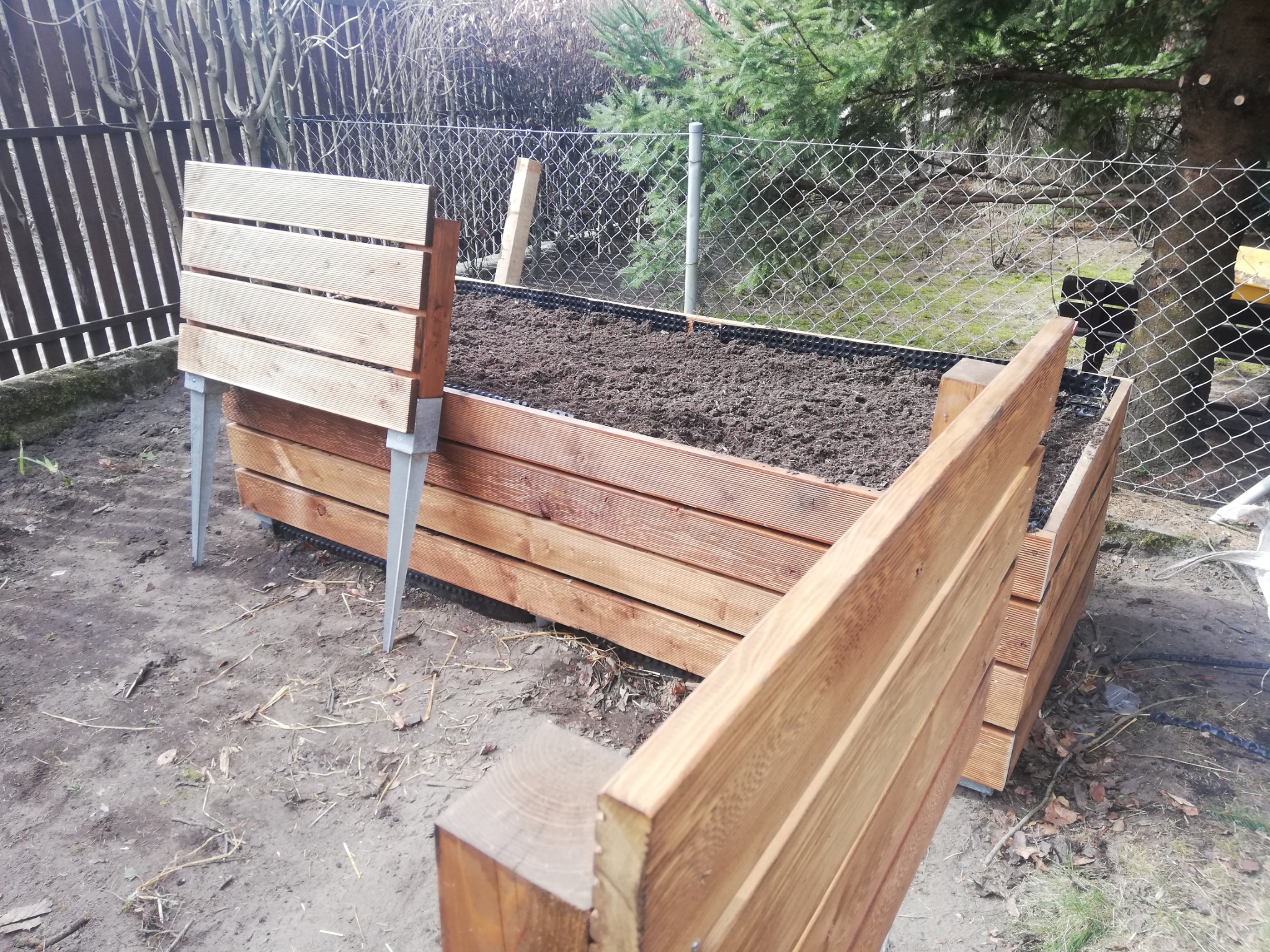 Podwyższona grządka warzywna z drewnianą obudową na metalowych nogach, wypełniona ziemią, widoczna siatka ogrodzeniowa w tle.
