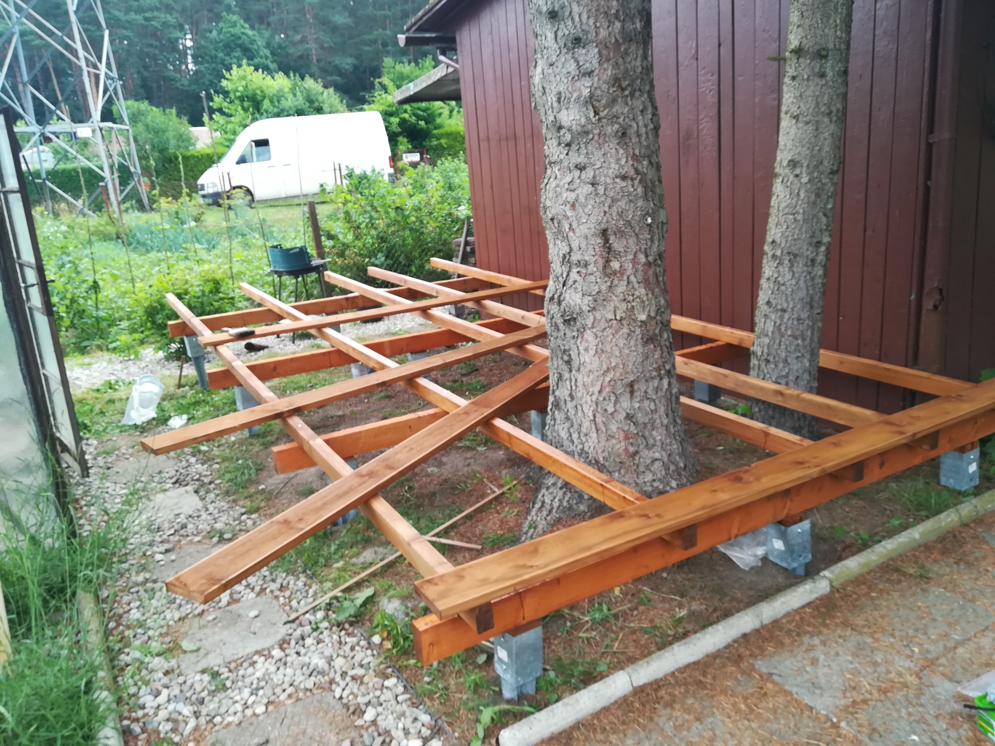Konstrukcja drewnianego podestu wokół dwóch drzew, wsparta na betonowych bloczkach, brązowy budynek w tle, biały van w oddali.