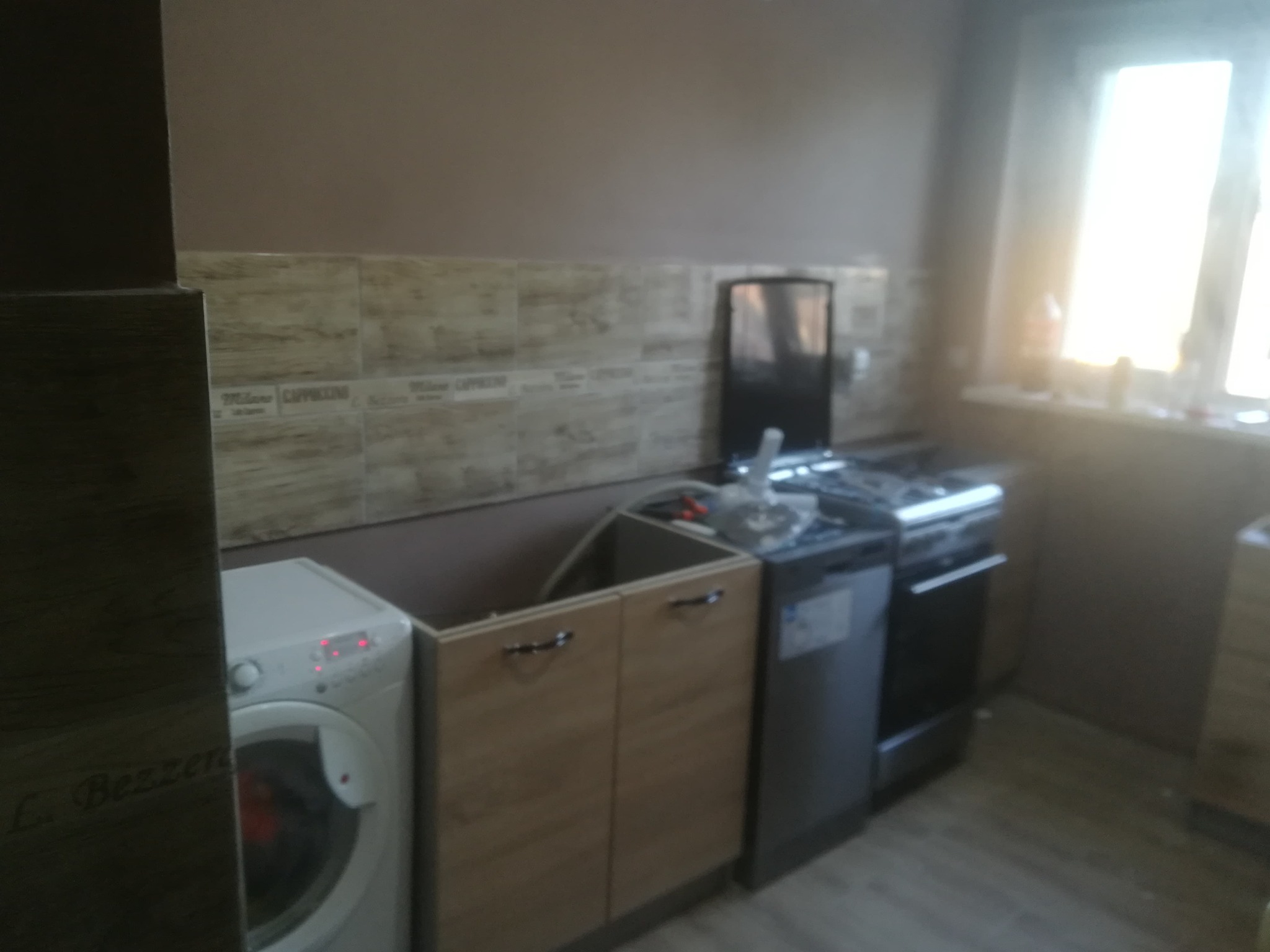 Łazienka w trakcie remontu: pralka, częściowo zamontowane szafki pod zlew, zmywarka i kuchenka, płytki ścienne z napisem 'Cappuccino', okno z silnym światłem dziennym.