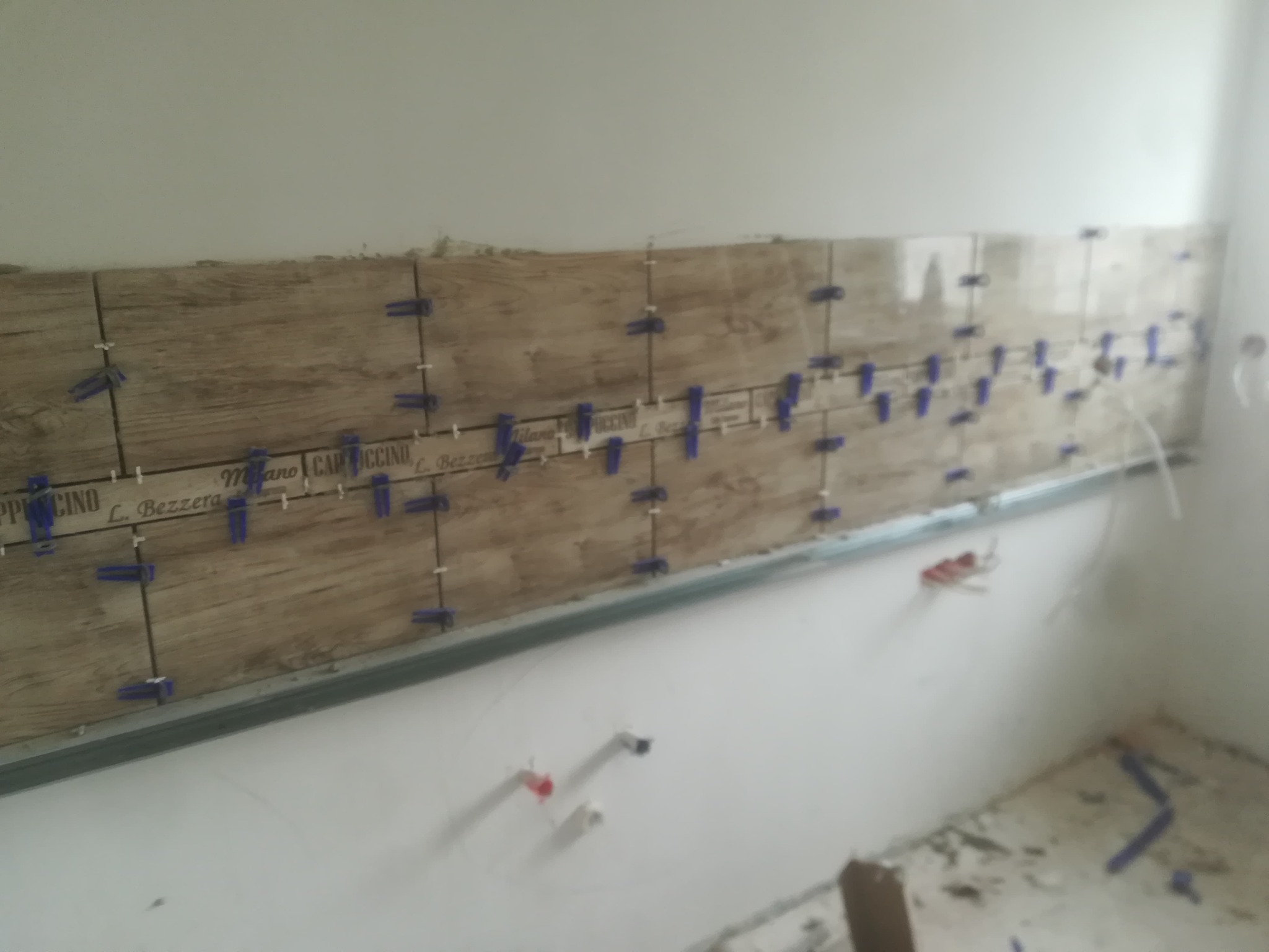 Ściana w trakcie układania płytek imitujących drewno z niebieskimi krzyżykami dystansowymi, widoczne przyłącza hydrauliczne na ścianie, fragment pomieszczenia w trakcie remontu.