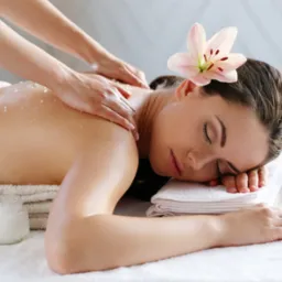 Kobieta z zamkniętymi oczami relaksuje się podczas masażu pleców z peelingiem solnym i lilią we włosach, na białym ręczniku leży słoiczek z kremem i bawełniany kompres ziołowy.