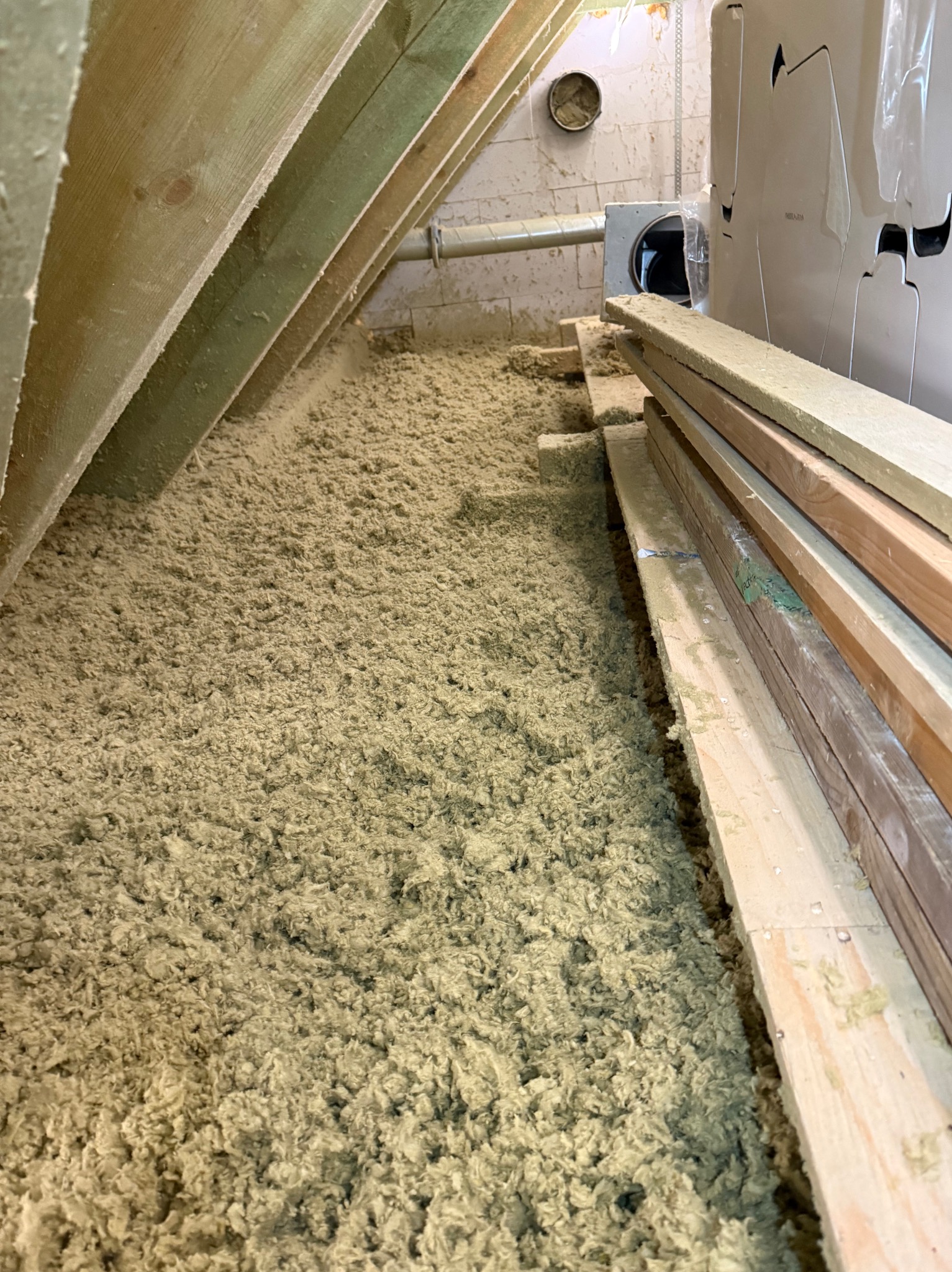 Poddasze z widoczną warstwą izolacji z wełny mineralnej, drewnianą konstrukcją więźby i fragmentem instalacji wentylacyjnej. Widoczne deski i elementy mocujące.