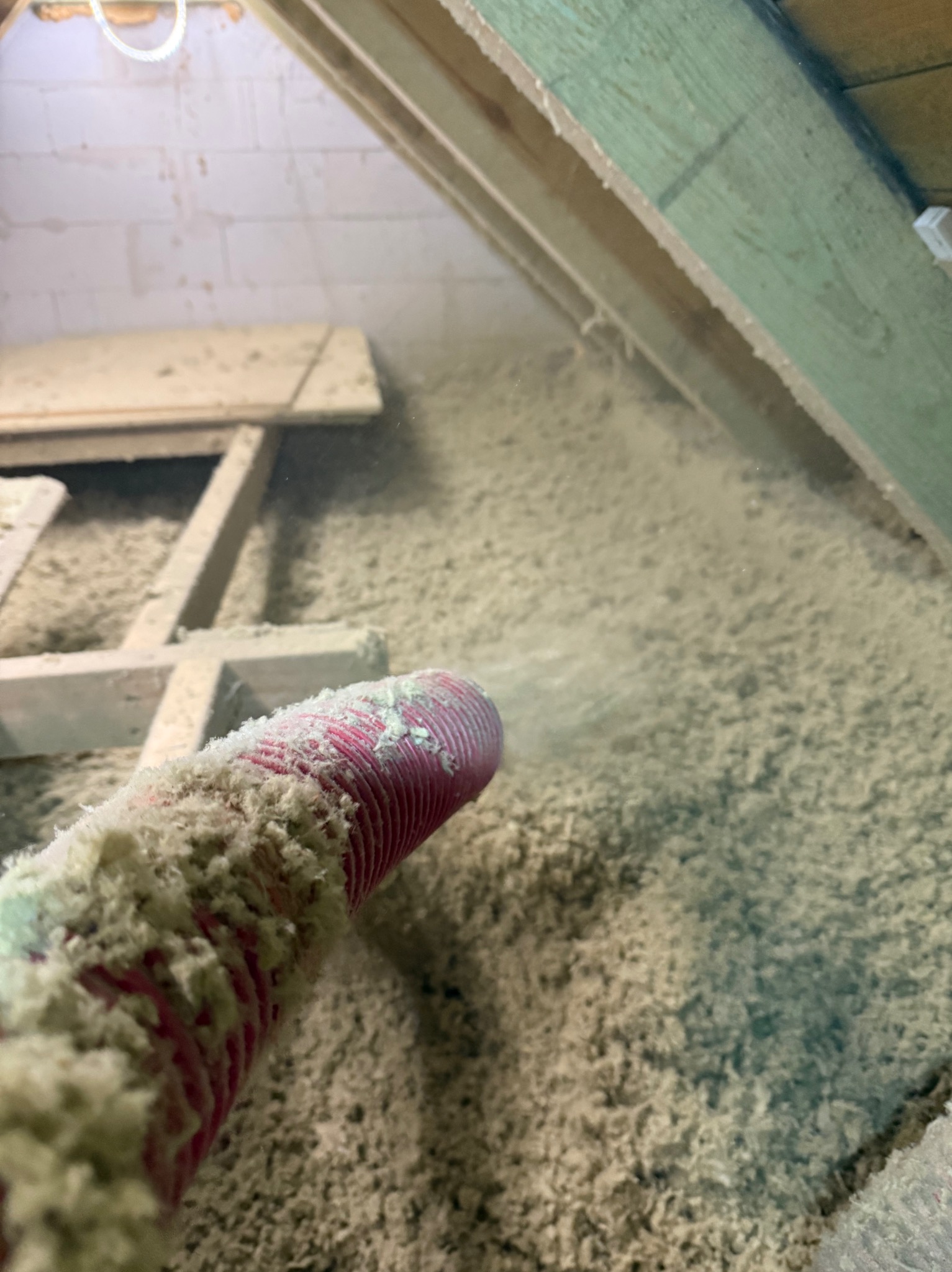 Aplikacja izolacji celulozowej w przestrzeni poddasza z użyciem węża. Widoczne drewniane elementy konstrukcyjne i biała izolacja.