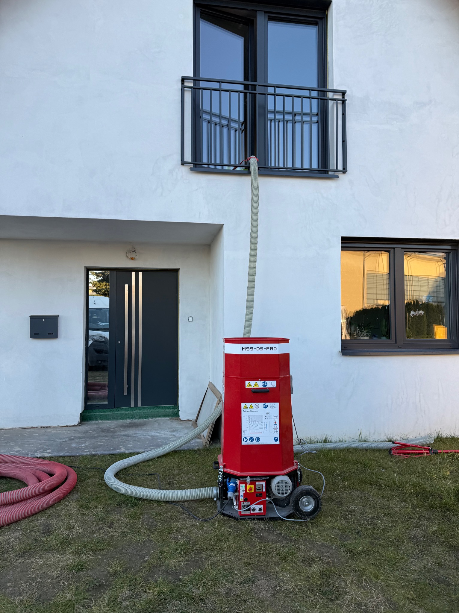 Czerwona maszyna M99-DS-PRO z podłączonym wężem do okna na białej elewacji budynku. Wokół trawa i zwinięte czerwone węże. Widok z dołu.