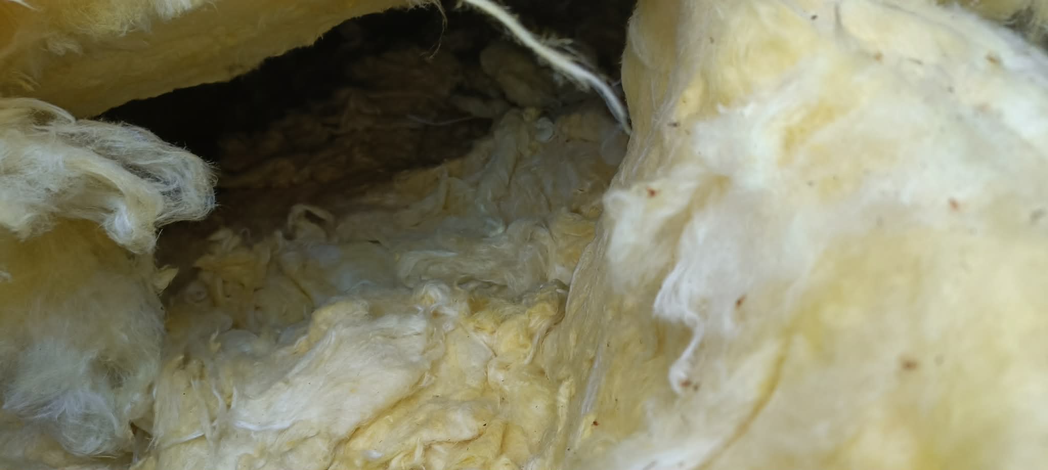 Żółta wełna mineralna wypełniająca przestrzeń poddasza, widoczna z bliska, z ciemnym obszarem w tle. Izolacja termiczna poddasza.