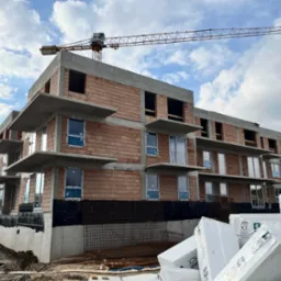 Budynek wielorodzinny w trakcie budowy: widoczne surowe ściany z cegły, zamontowane okna z folią ochronną, betonowe balkony, żuraw budowlany w tle oraz materiały izolacyjne na pierwszym planie.