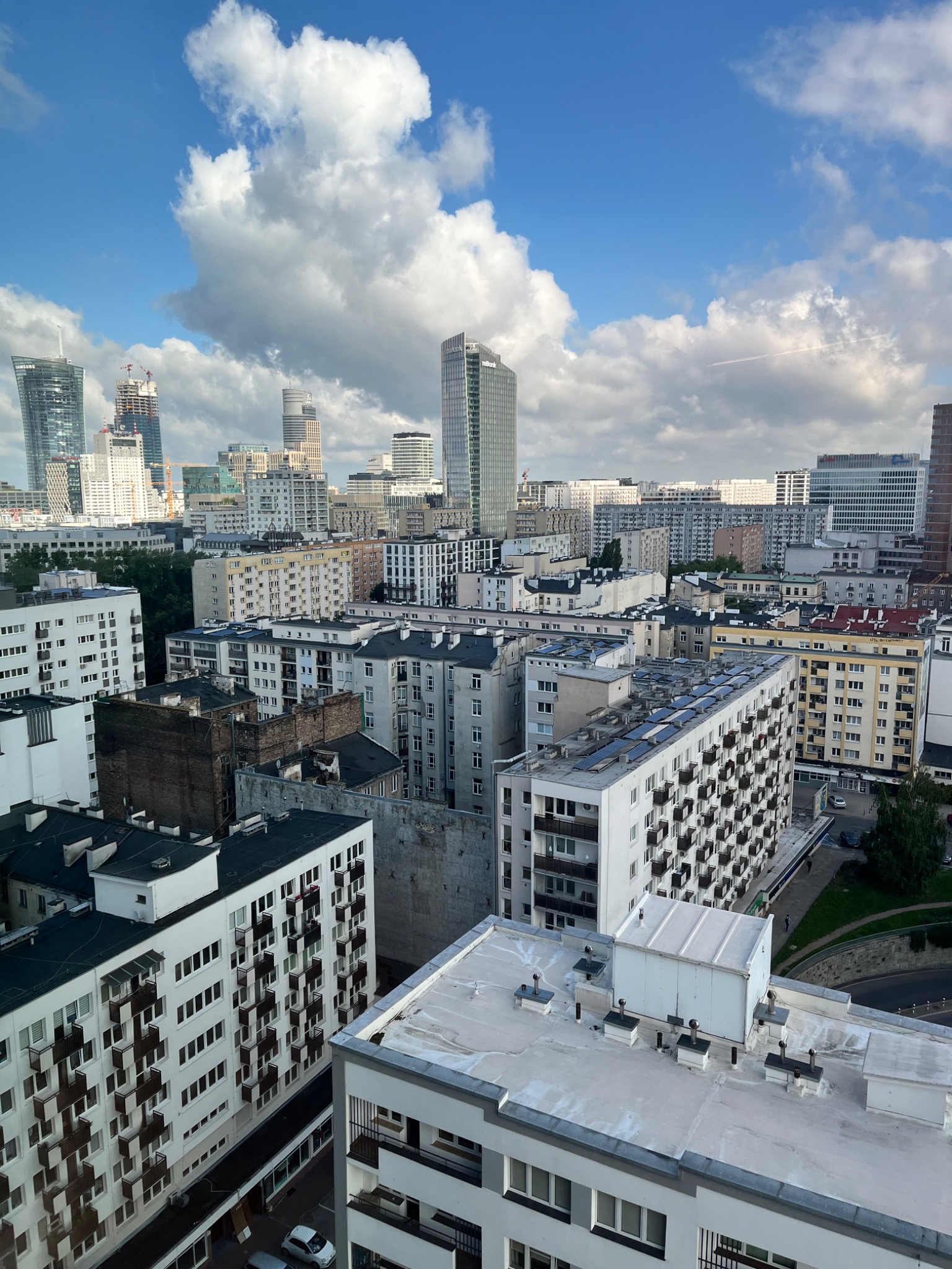 Panorama Warszawy z widokiem na budynki mieszkalne, biurowce i chmury na błękitnym niebie. Na dachu jednego z budynków widoczne panele słoneczne.