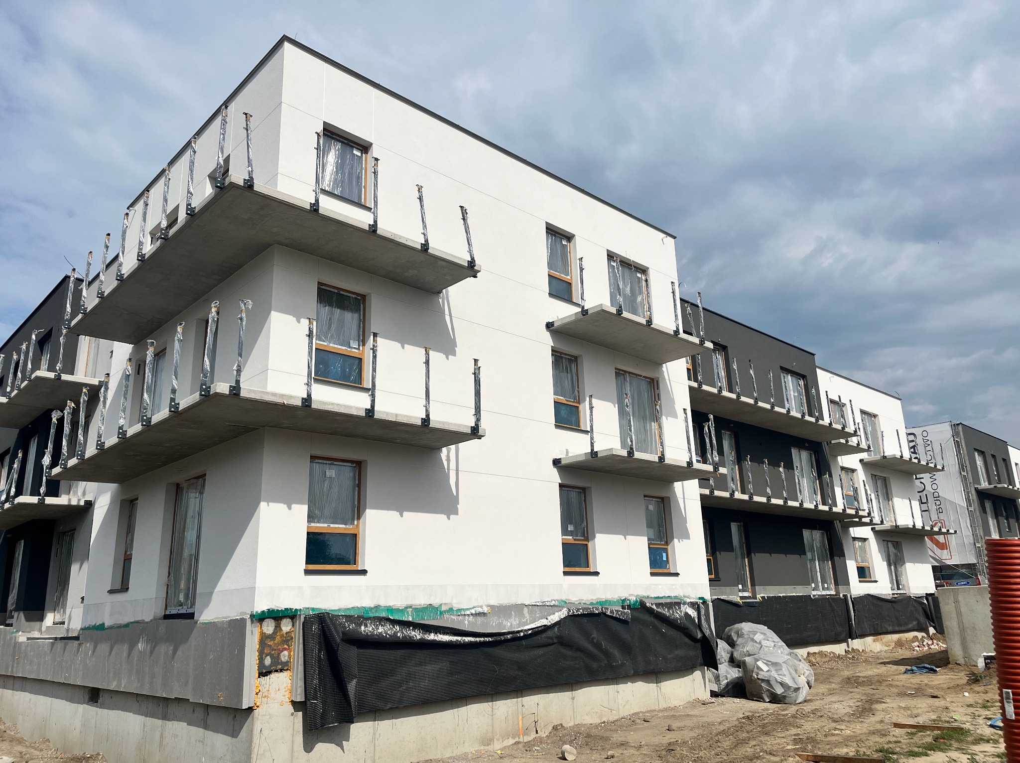 Nowoczesny budynek wielorodzinny w trakcie wykańczania elewacji, z widocznymi balkonami zabezpieczonymi folią, czarna membrana hydroizolacyjna u podstawy i materiały budowlane na placu budowy.