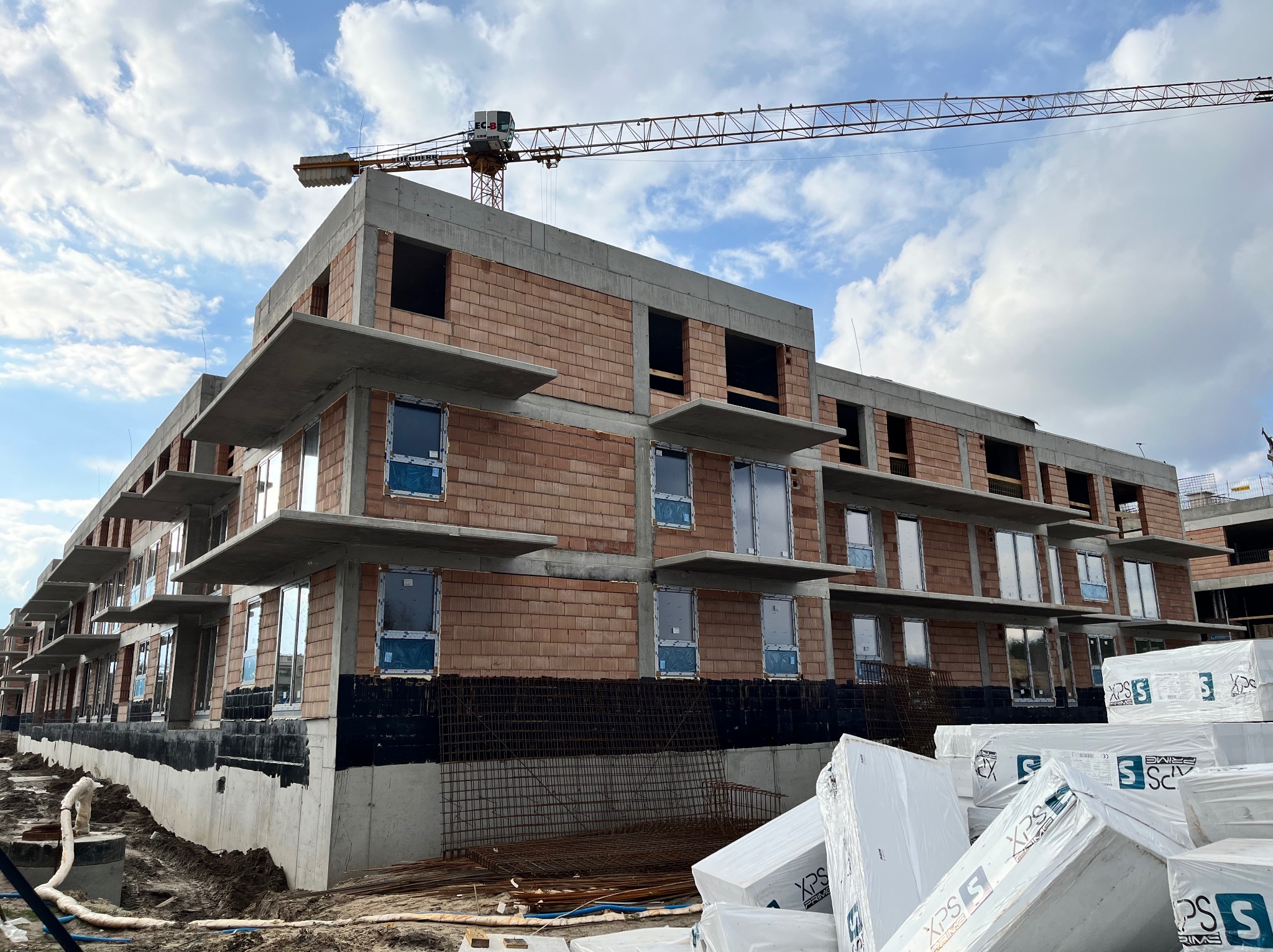 Budynek wielorodzinny w trakcie budowy: widoczne surowe ściany z cegły, zamontowane okna z folią ochronną, betonowe balkony, żuraw budowlany w tle oraz materiały izolacyjne na pierwszym planie.