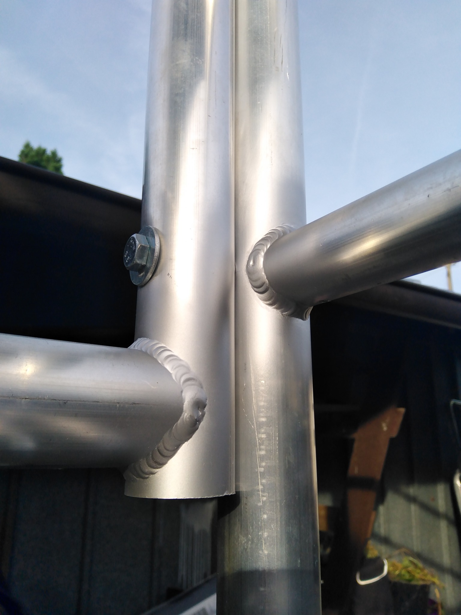 Szczegółowe zbliżenie na spawane połączenie rur aluminiowych, widoczne ślady spawania i śruba mocująca, na tle fragmentu budynku.