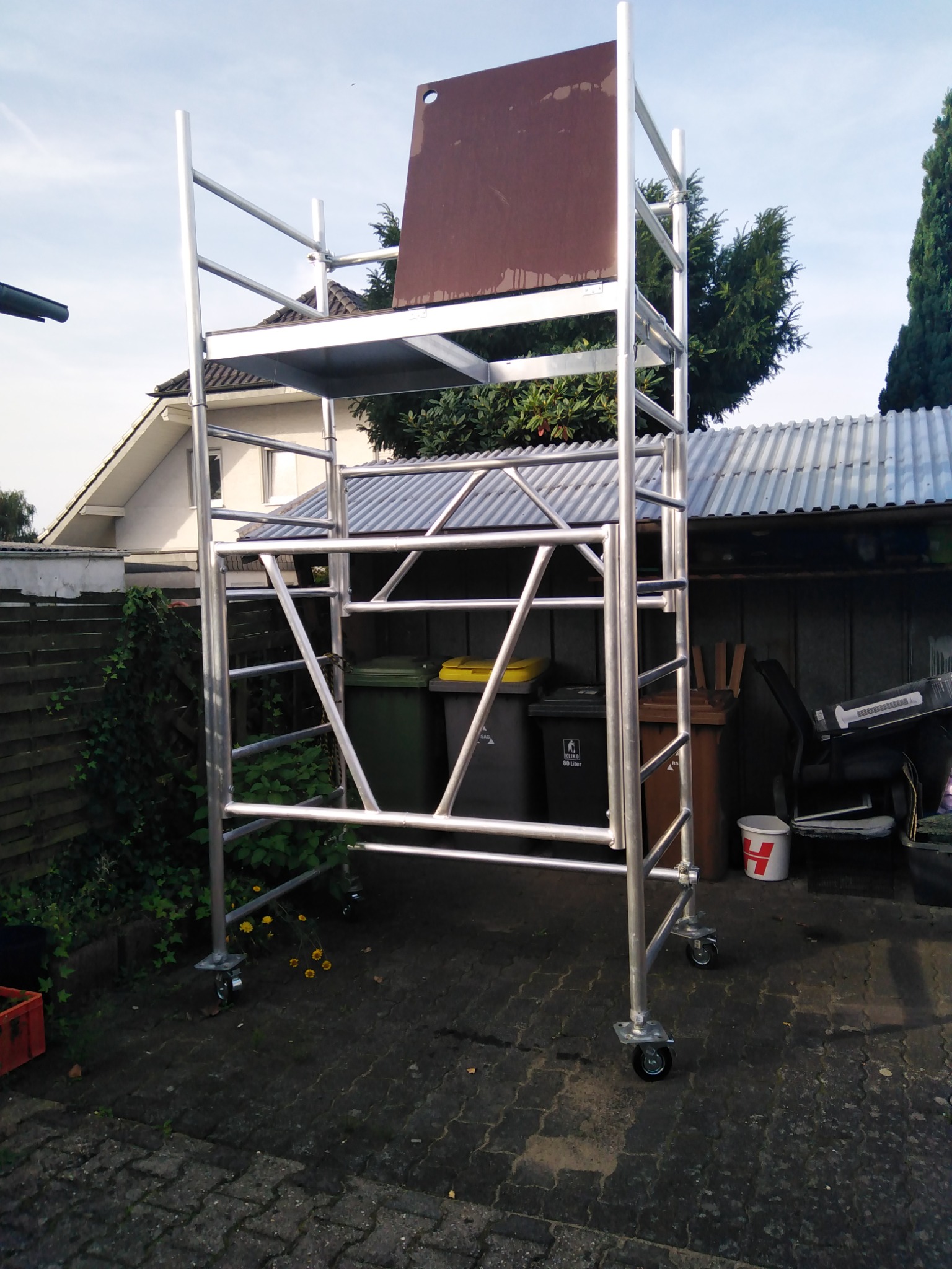 Aluminiowe rusztowanie na kółkach z brązową platformą ochronną na tle domu i zieleni, stojące na nierównym bruku.