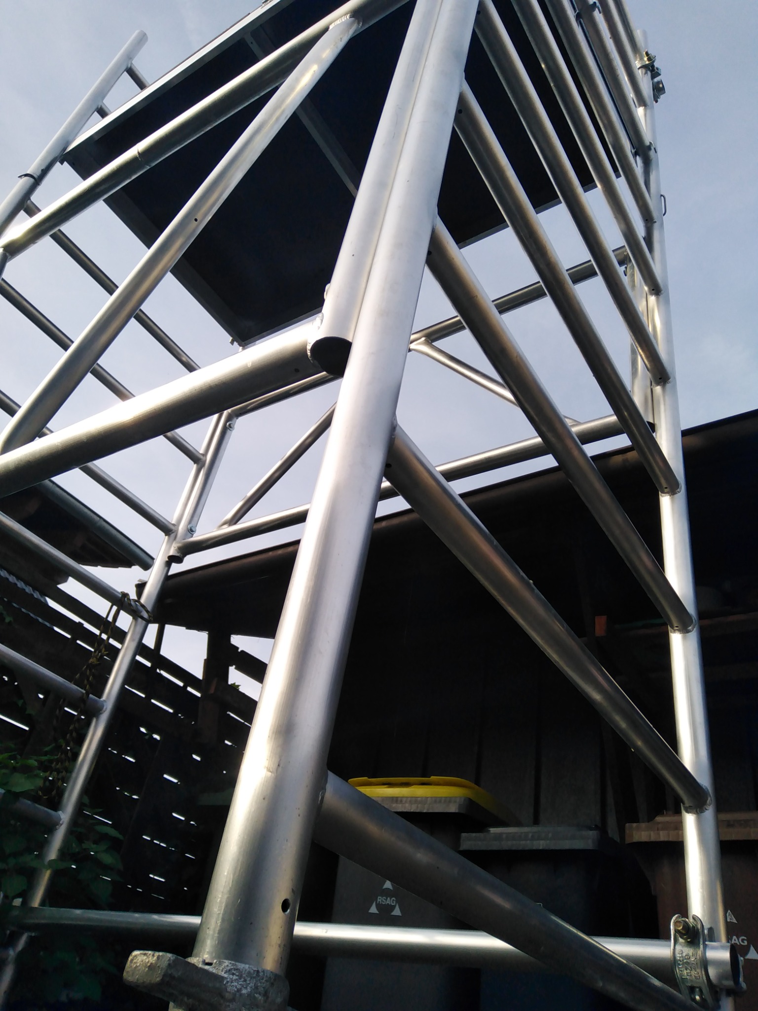 Metalowa konstrukcja rusztowania z widocznymi spawami i elementami mocującymi, ustawiona przy budynku z drewnianą elewacją.