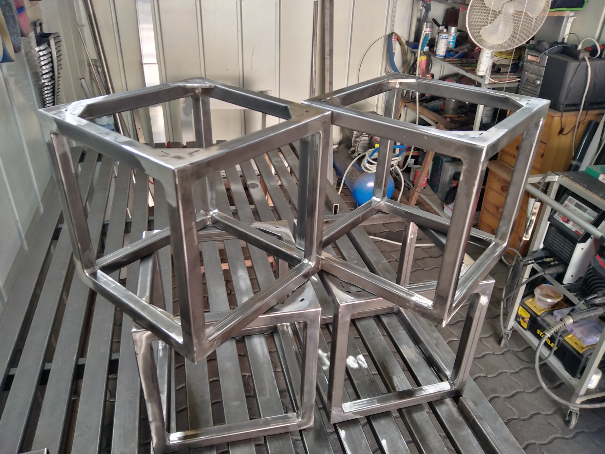 Surowe, spawane metalowe ramy w kształcie sześcianów, ustawione na metalowej kratownicy w warsztacie, widoczne ślady spawania.