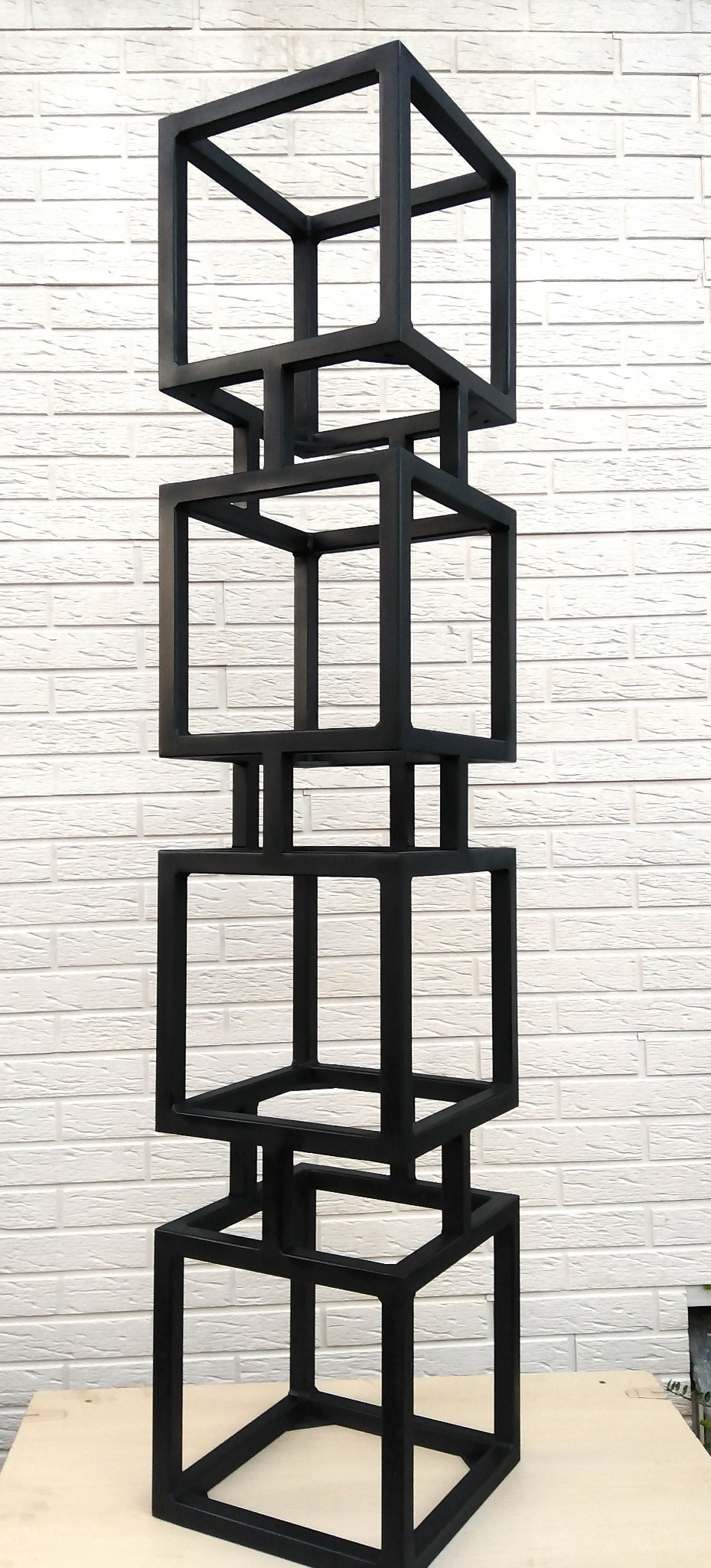 Czarna, metalowa konstrukcja składająca się z połączonych kwadratowych ram, ustawiona na jasnym tle ściany z cegieł.