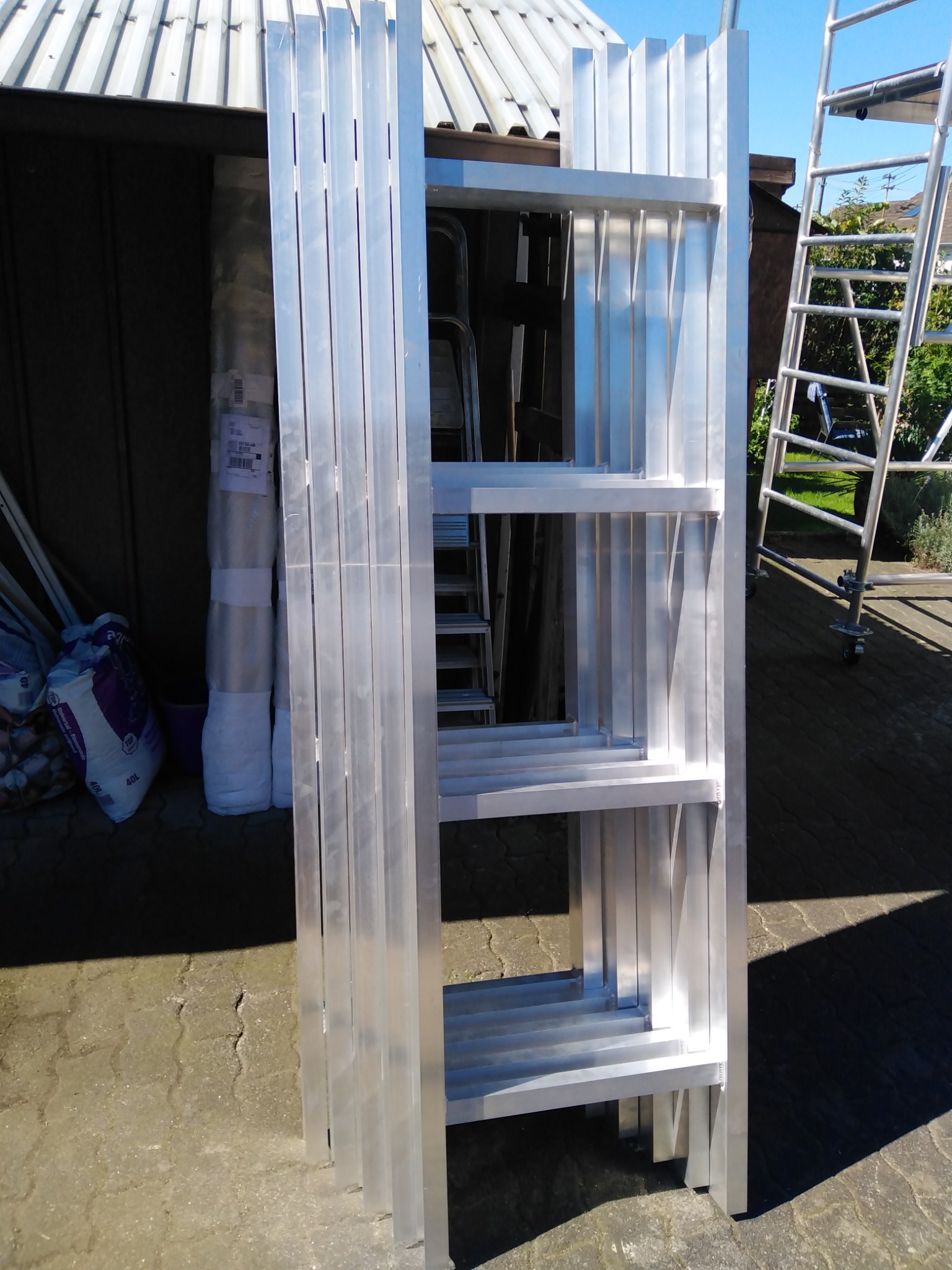 Stos elementów konstrukcji schodów metalowych z profili aluminiowych, opartych o ścianę budynku gospodarczego, w tle widoczna drabina rusztowania.