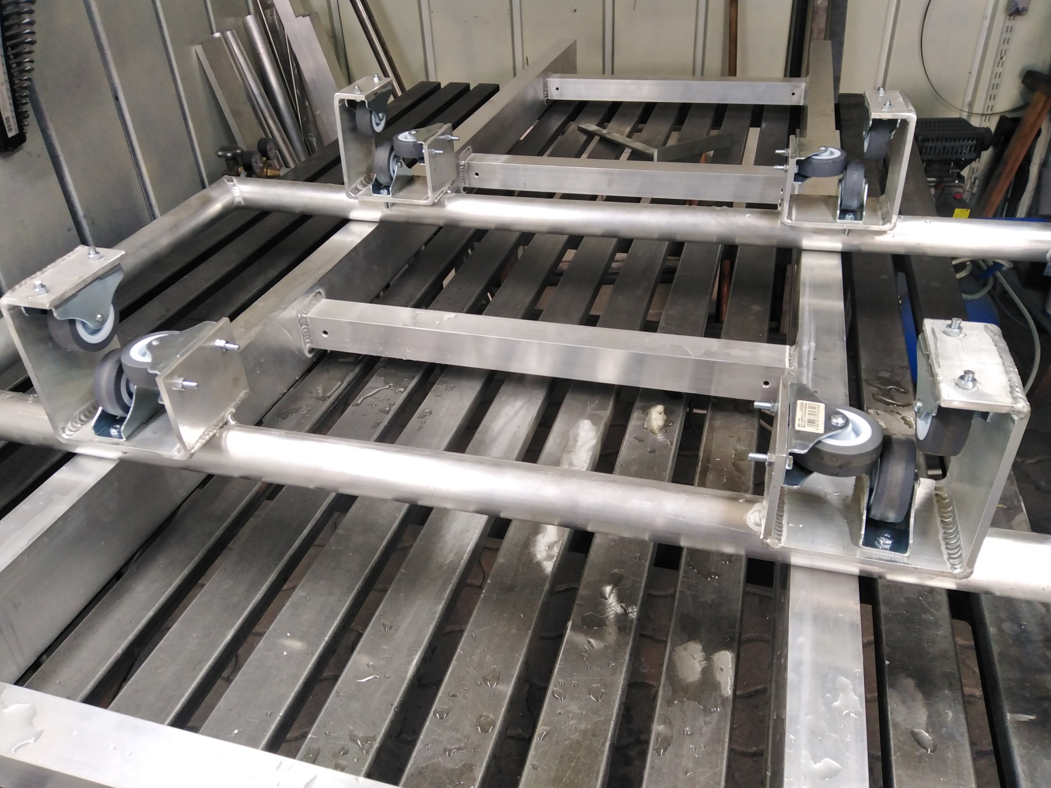 Stalowa rama z zamontowanymi kółkami transportowymi, spawana na metalowym stole warsztatowym, widoczne ślady obróbki i spawania.