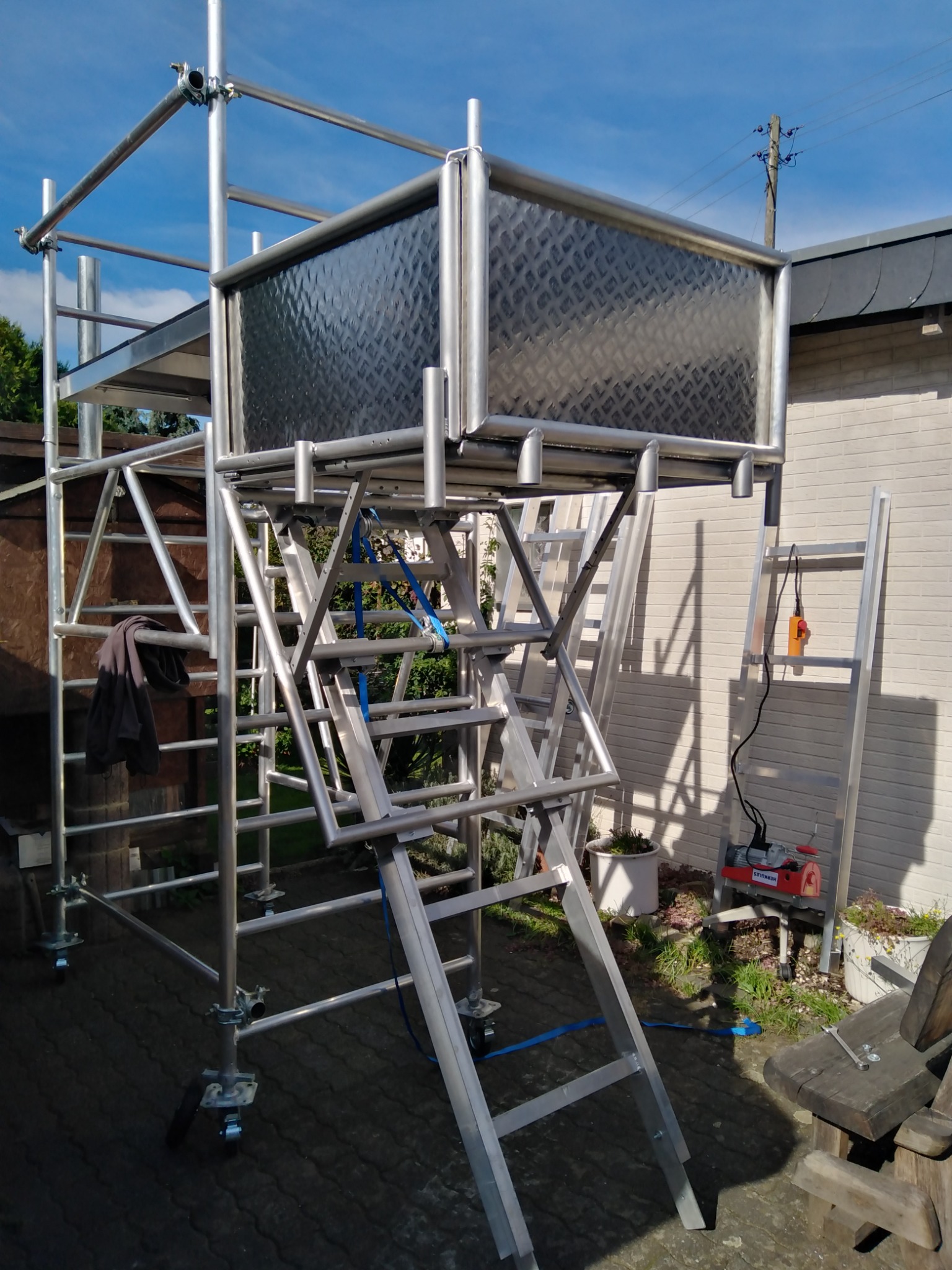 Mobilny, metalowy pomost roboczy na kółkach z drabinkami, z platformą roboczą z blachy ryflowanej, stojący na brukowanym podwórku przy ścianie budynku.