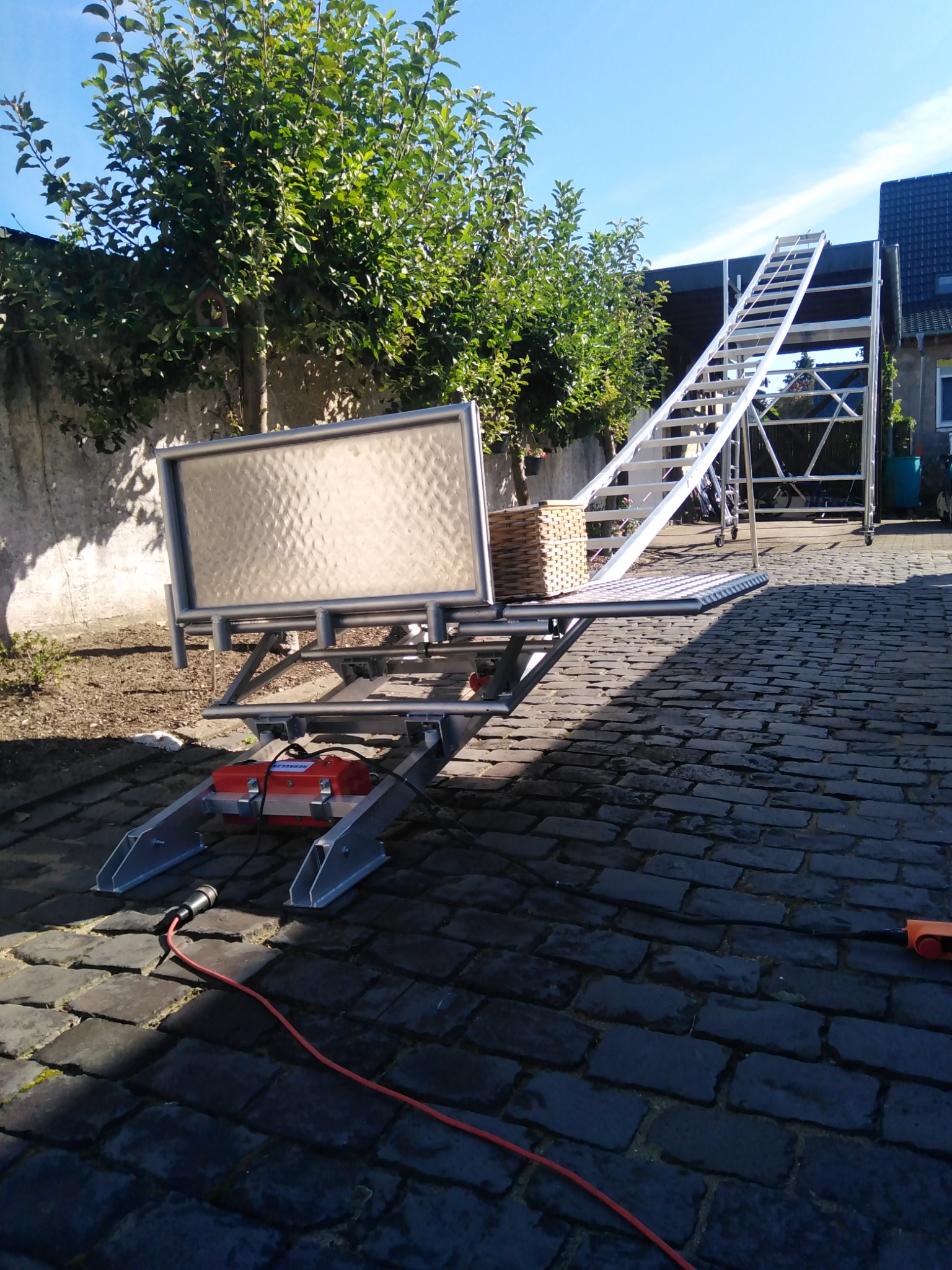 Mobilna winda budowlana z platformą i schodami ustawiona na brukowanym podwórku, z koszem na platformie i widocznym przewodem zasilającym.