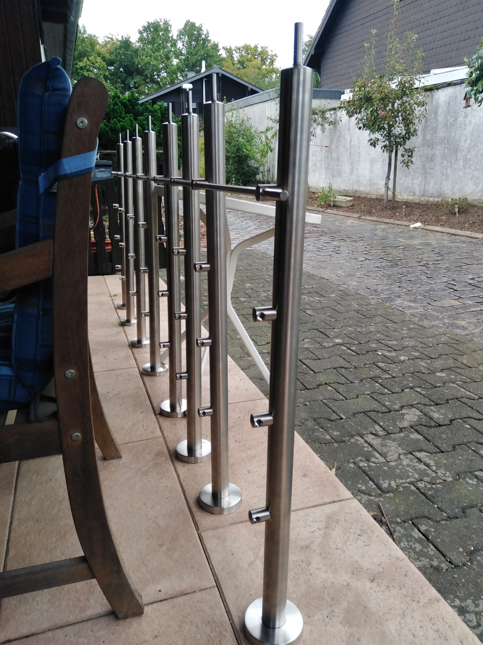 Rząd słupków balustrady ze stali nierdzewnej, zamontowanych na tarasie, widoczny fragment drewnianego krzesła i kostki brukowej.