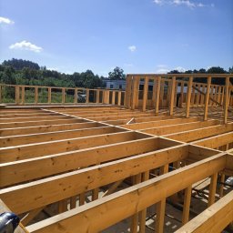 Comfort studio - Drewniana konstrukcja szkieletowa domu w trakcie budowy, widoczne belki stropowe i słupy nośne, narzędzie akumulatorowe na pierwszym planie, w tle zieleń i błękitne niebo.