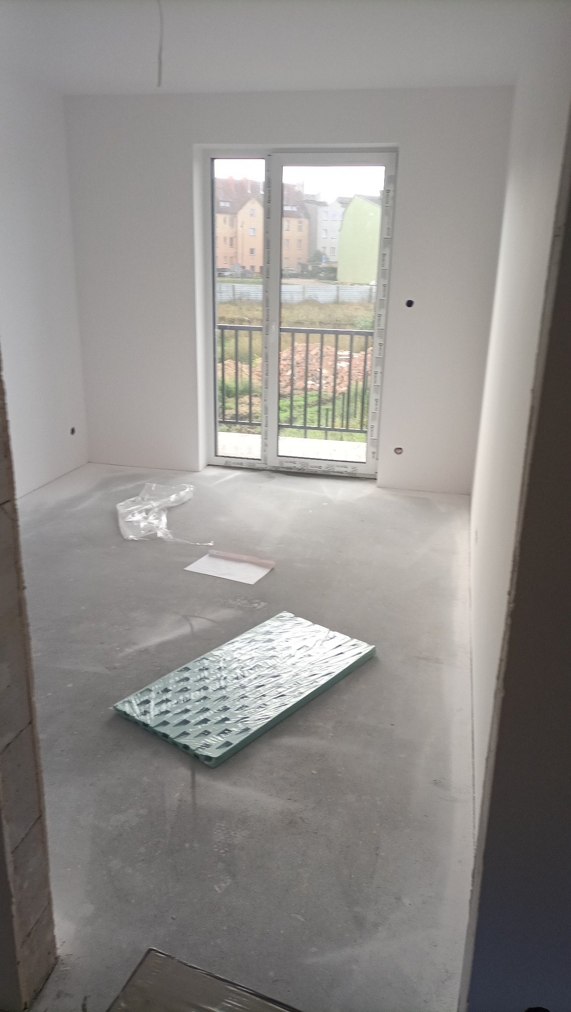 Surowe wnętrze pokoju z przygotowanym podłożem, paczka zielonych płyt izolacyjnych leży na szarym, betonowym podłożu, ściany pomalowane na biało, widok z okna na budynki mieszkalne.