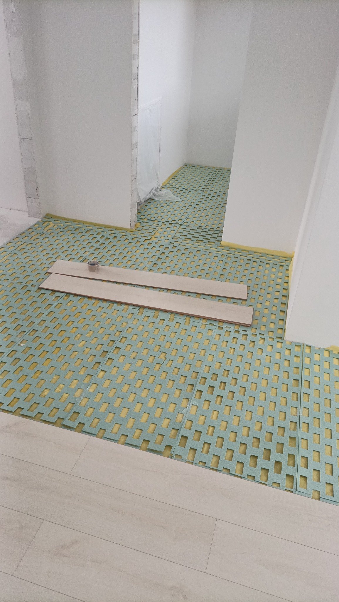 Przygotowanie podłoża pod parkiet: zielona mata z otworami rozłożona na podłodze, widoczne dwie deski parkietowe i pojemnik z klejem, fragment ułożonej podłogi w rogu kadru.