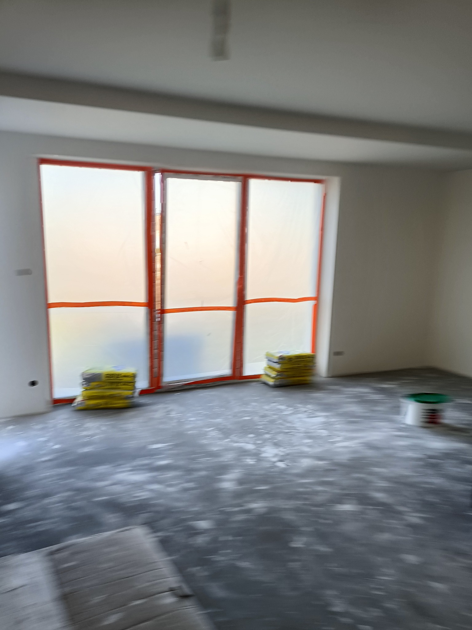 Przygotowanie pokoju do prac wykończeniowych: okna zabezpieczone folią, worki z zaprawą i wiadro farby w pustym pomieszczeniu z szarą posadzką.