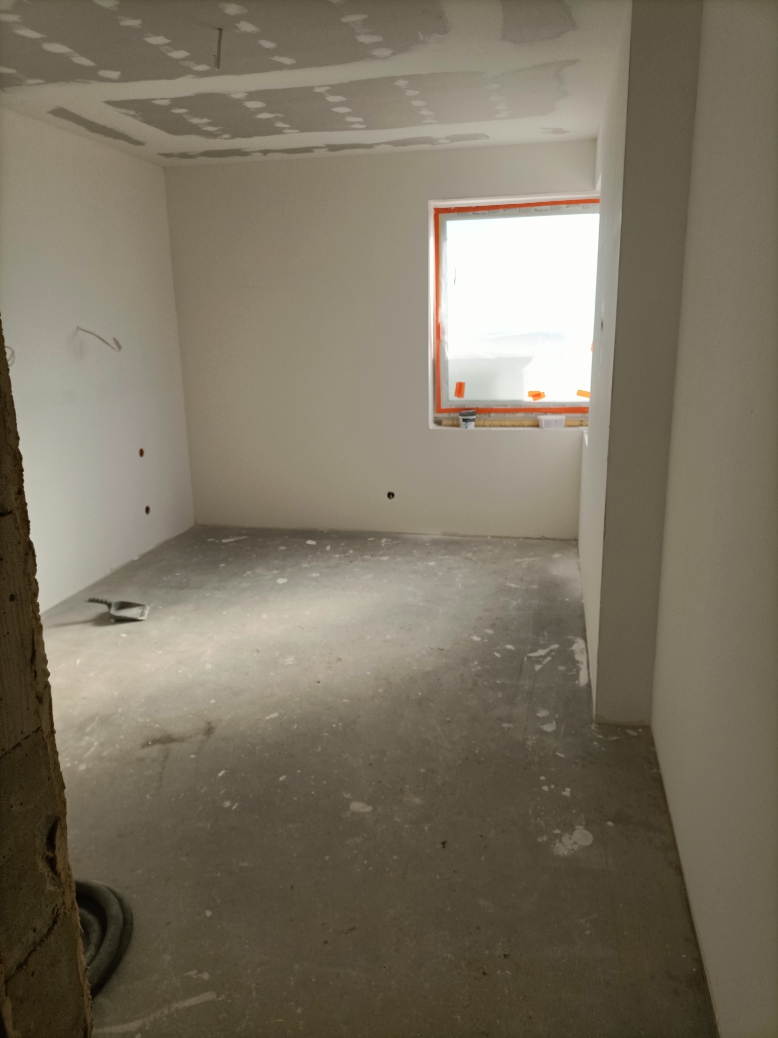 Pokój w trakcie remontu: surowy beton na podłodze, ściany przygotowane do malowania, częściowo wykończony sufit z płyt karton gips, okno zabezpieczone folią ochronną.