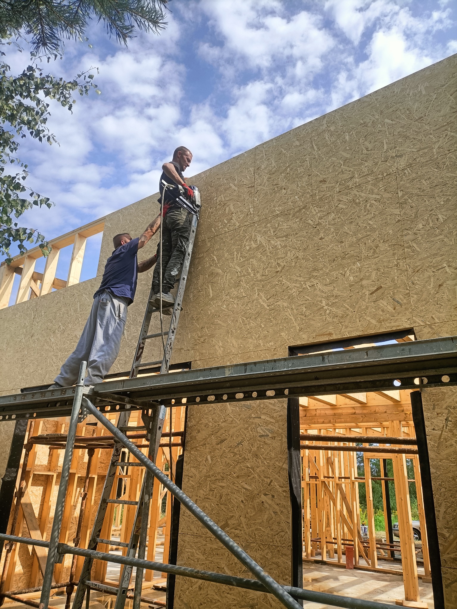 Dwóch mężczyzn montuje płyty OSB na ścianie szkieletowej budynku, jeden na rusztowaniu, drugi na drabinie, używając narzędzia pneumatycznego, widoczne niebo z chmurami.