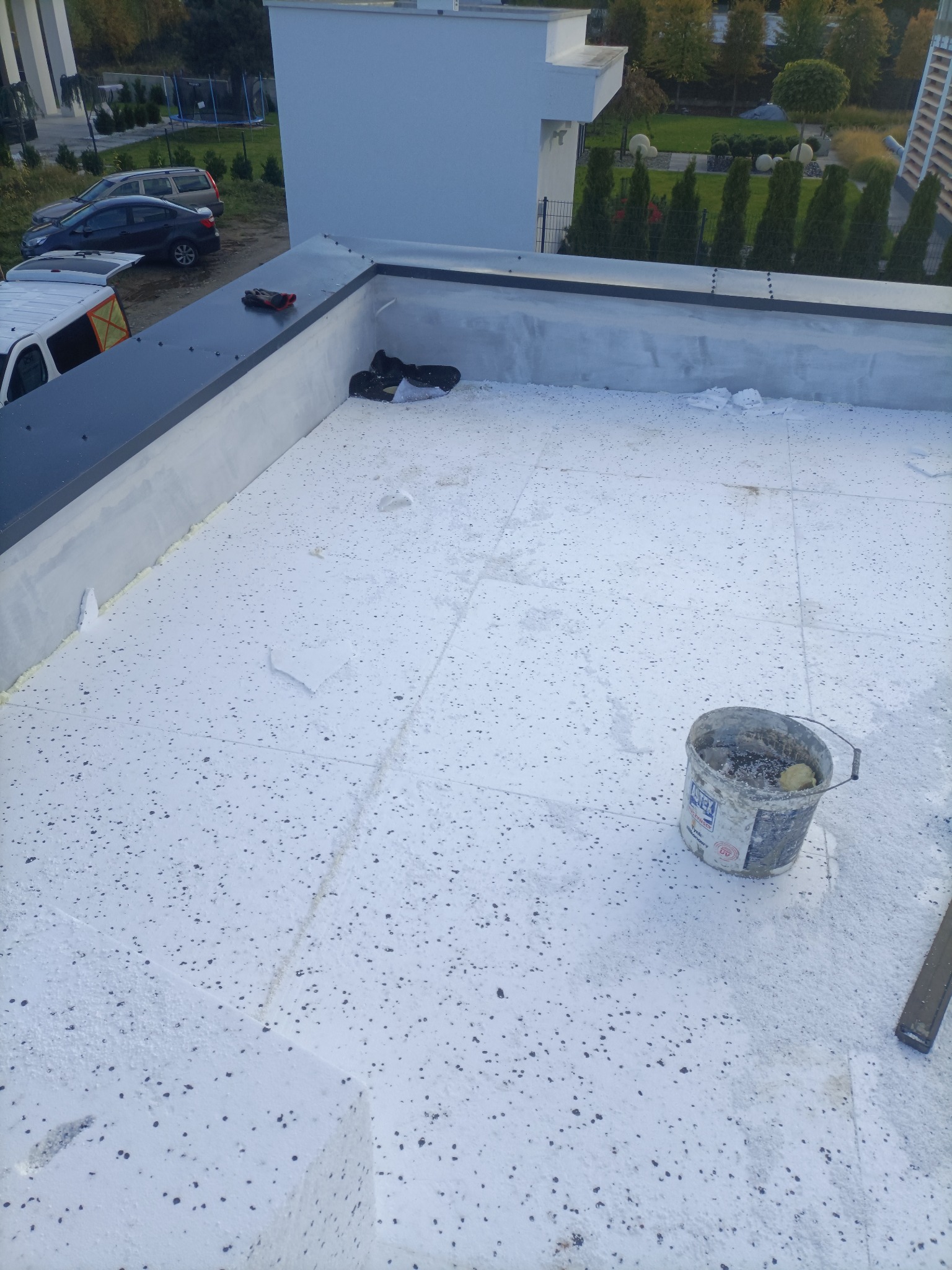 Biała powierzchnia dachu pokryta styropianem z widocznym wiadrem po kleju i fragmentami styropianu, w tle budynek i zieleń.
