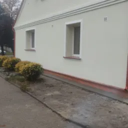 Jasna elewacja domu jednorodzinnego po renowacji, z widocznymi oknami w białych ramach i pomarańczowym obramowaniem u dołu, obok chodnik z kostki brukowej i krzewy.