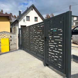 Marcin Gryz - Nowoczesna, przesuwna brama wjazdowa z ażurowym wzorem, pomalowana na grafitowo. Widoczny fragment domu z białą elewacją i żółta skrzynka gazowa.