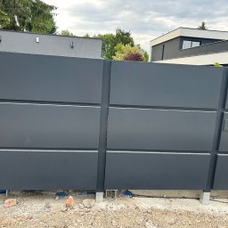 Marcin Gryz - Nowoczesna, przesuwna brama wjazdowa w kolorze grafitowym z widoczną skrzynką na listy i instalacją elektryczną w ziemi. Tło stanowią budynki mieszkalne i zieleń.