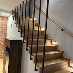 Marcin Gryz - Drewniane schody z czarną, metalową balustradą w nowoczesnym wnętrzu. Stopnie oświetlone punktowymi lampami. Widoczna część ściany z cegły i drewniana komoda.