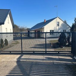 Marcin Gryz - Nowoczesna, przesuwna brama wjazdowa w kolorze grafitowym, z pionowymi szczeblami, na tle jasnego domu z ciemnym dachem i błękitnego nieba w słoneczny dzień.