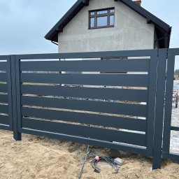 Rzemiosło - Nowoczesna, przesuwna brama wjazdowa z poziomymi panelami w kolorze grafitowym, na tle domu w trakcie budowy. Widoczne przewody elektryczne przy fundamencie.